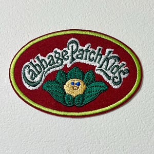 Puede incluir: Un parche bordado ovalado rojo con texto blanco que dice "Cabbage Patch Kids" y un niño de la col verde con cara amarilla y ojos azules.