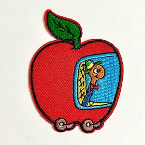 May include: An embroidered patch shaped like a red apple with a green leaf on top. The apple features a blue window showing a tan worm wearing a green hat and a red bowtie. Two small brown wheels are attached to the bottom of the apple shape.