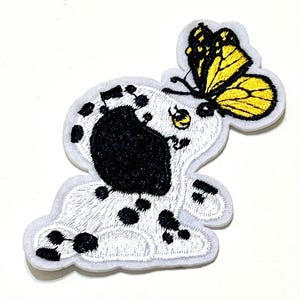 Puede incluir: Parche bordado con un perro dálmata blanco y negro con una gran oreja negra y una mariposa amarilla y negra. El parche tiene un borde blanco y está diseñado para su aplicación en ropa o accesorios.