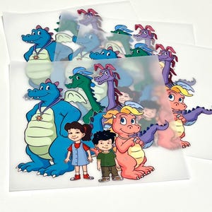 Puede incluir: Un conjunto de hojas de transferencia translúcidas con coloridas ilustraciones de dibujos animados de un dragón azul con colgante, un dragón verde, un dragón morado, un dragón rosa y dos niños de pie juntos, impresas en una película transparente.