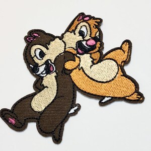 May include: Embroidered patch featuring Chip and Dale, the Disney chipmunk duo. The patch shows the characters in a playful embrace, with brown, tan, and pink colors. The patch has a textured appearance and a dark brown border.