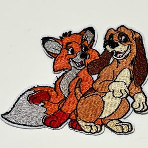 May include: Embroidered patch featuring two cartoon characters: an orange fox and a brown hound dog. The fox has a white chest and gray accents, while the dog has floppy ears and a friendly expression. The patch has a white border.