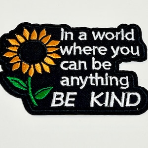 May include: Embroidered patch with a black background, featuring a sunflower and the text "In a world where you can be anything, BE KIND" in white lettering. The sunflower has yellow petals and a dark center.