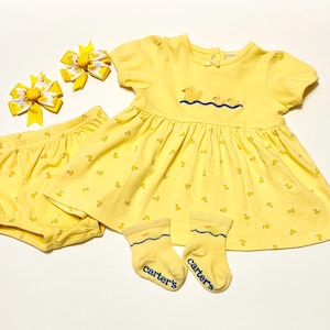May include: A yellow baby clothing set featuring a dress, bloomers, socks, and two hair bows. The dress and bloomers are covered in small duck prints. The dress has an embroidered duck design on the chest. The socks have the brand name "Carter's" on them.