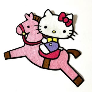 Könnte beinhalten: Gestickter Aufnäher mit einer Cartoon-Katze mit roter Schleife, die ein rosa Pferd reitet. Die Katze trägt ein gelbes und lila Outfit. Das Pferd hat braune Hufe und eine braune Mähne. Der Aufnäher ist mit einem weißen Hintergrund gestaltet.