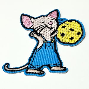 Può includere: Toppa ricamata raffigurante un topo dei cartoni animati con una tuta blu e che tiene in mano un grande biscotto giallo con gocce di cioccolato nere. Il topo ha orecchie rosa e un viso bianco, con contorni neri. La toppa ha un bordo blu.