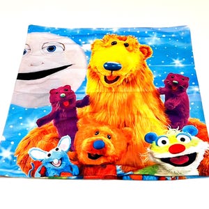 Könnte beinhalten: Ein farbenfrohes, quadratisches Stoffstück mit Cartoonfiguren. Das Design umfasst einen großen gelben Bären, kleinere orangefarbene und violette Bären, eine blaue Maus, einen lächelnden Mond und einen weißen Hintergrund mit blauen Schneeflocken.