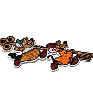 May include: Embroidered patch featuring two cartoon characters, a bear and a dog, each holding a musical instrument. The bear wears a green hat and the dog an orange shirt. The patch has a white border.