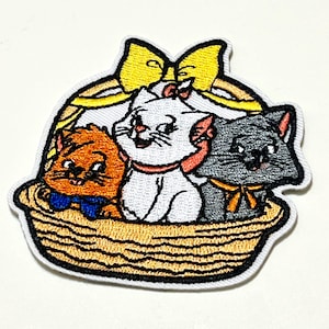 以下が含まれることがあります： 織りバスケットに入った3匹の漫画の子猫が描かれた刺繍パッチ。子猫はオレンジ、白、グレーで、黄色とピンクのアクセントが付いています。大きな黄色のリボンがバスケットの上にあります。