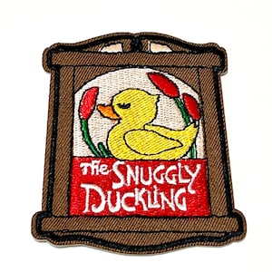 Op de afbeelding: Geborduurde patch met een gele eend in een bruine lijst. De eend is omgeven door rode en groene details, met de tekst "The Snuggly Duckling" in het wit op een rode banner. De patch heeft een bruine rand.