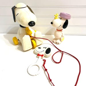 Op de afbeelding: Vintage Snoopy-speelgoed in gebroken wit, zwart, geel en rood. Het speelgoed omvat een Snoopy om aan te trekken, een Snoopy-beeldje met een hoed en een Snoopy-gezicht met een rode vlinderdas. Een rood touwtje verbindt het speelgoed.