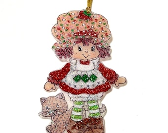 May include: A glittery hanging ornament featuring a character with a wide-brimmed hat decorated with strawberries, wearing a red dress with a white apron, green and white striped stockings, and brown shoes, accompanied by a small spotted cat.