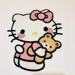 Peut inclure: Patch thermocollant brodé représentant Hello Kitty dans une robe rose tenant un ours en peluche brun.