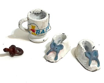 May include: A collection of miniature baby items, including a white mug with a "BABY" banner and a cartoon bear, two white baby shoes with blue bows, and a brown pacifier. The items are arranged on a white surface.