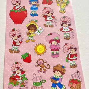 May include: A sheet of stickers featuring Strawberry Shortcake and her friends. The stickers are on a pink background with white polka dots. The stickers feature various characters, including Strawberry Shortcake, Raspberry Tart, Orange Blossom, Lemon Meringue, Blueberry Muffin, and Huckleberry Pie. The stickers also feature a variety of animals, including a cat, a monkey, and a bird.