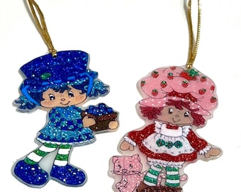 May include: Two vintage-style ornaments featuring cartoon characters. One is a blue character holding a basket of blueberries, and the other is a character in a red dress and a pink hat with a pink cat. Both have gold hanging strings.