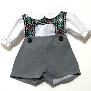 Può includere: Una camicia bianca a maniche lunghe con colletto arrotondato abbinata a pantaloncini grigi con bretelle. Le bretelle sono decorate con un motivo geometrico a rombi nero, rosso, blu e bianco. Due bottoni scuri fissano le bretelle ai pantaloncini.