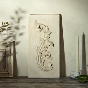 Peut inclure: Plaque murale rectangulaire blanc cassé avec un motif floral et de feuilles en relief. La pièce décorative est placée contre un mur neutre, avec une bougie et un objet encadré en arrière-plan. La plaque mesure environ 30 cm de large et 61 cm de haut.