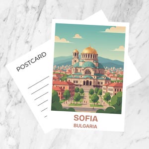 May include: A postcard featuring an illustration of a cathedral with gold domes and a cityscape. The postcard has the text "SOFIA BULGARIA" at the bottom. A blank postcard is behind the illustrated postcard. The background is a marble surface.