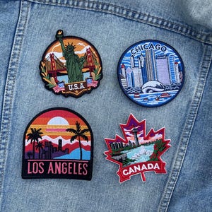 May include: Four embroidered patches on a denim jacket. Patches feature the Statue of Liberty, Chicago skyline, Los Angeles sunset, and a Canadian maple leaf with text that reads "U.S.A", "CHICAGO", "LOS ANGELES", and "CANADA".