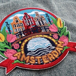 Puede incluir: Parche bordado con una colorida escena de &Aacute;msterdam con casas canal, un puente y tulipanes. El parche tiene un borde rojo y una pancarta con el nombre de la ciudad. El dise&ntilde;o incluye colores azul, naranja, rojo y verde.
