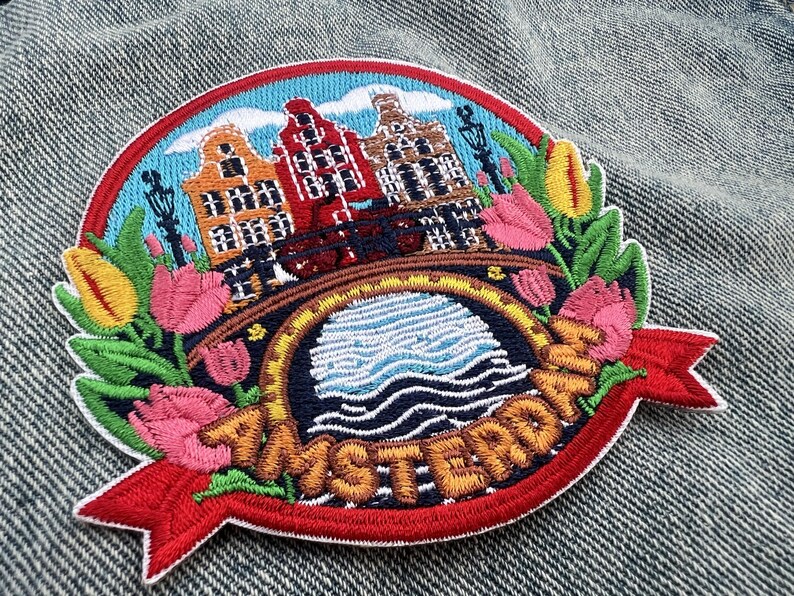 Puede incluir: Parche bordado con una colorida escena de &Aacute;msterdam con casas canal, un puente y tulipanes. El parche tiene un borde rojo y una pancarta con el nombre de la ciudad. El dise&ntilde;o incluye colores azul, naranja, rojo y verde.