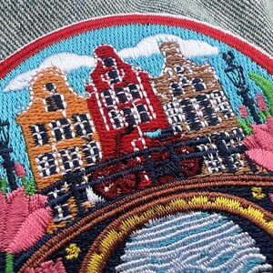 Puede incluir: Parche bordado con un colorido paisaje urbano con tres casas de canal en naranja, rojo y marr&oacute;n. El dise&ntilde;o incluye un puente, tulipanes y un cielo azul con nubes, todo enmarcado por un borde rojo y azul. Ideal para ropa o accesorios.