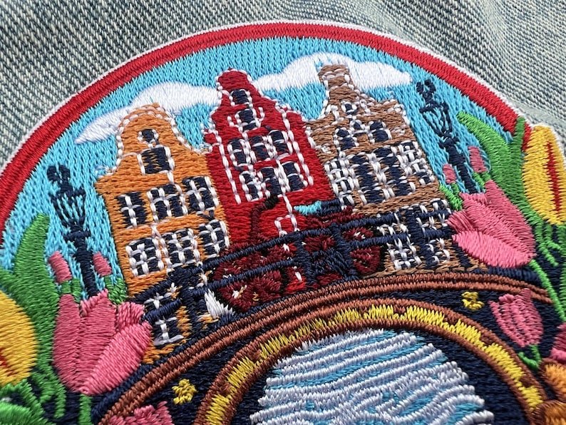 Puede incluir: Parche bordado con un colorido paisaje urbano con tres casas de canal en naranja, rojo y marr&oacute;n. El dise&ntilde;o incluye un puente, tulipanes y un cielo azul con nubes, todo enmarcado por un borde rojo y azul. Ideal para ropa o accesorios.