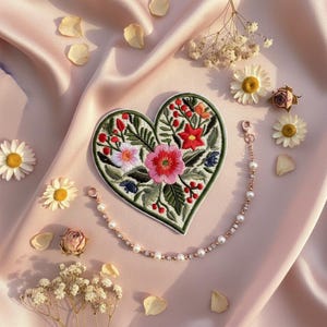 Könnte beinhalten: Ein herzförmiger, gestickter Aufnäher mit einem Blumendesign in Rot, Rosa und Weiß, umgeben von grünen Blättern. Eine zarte Perlen- und Goldkette ist befestigt. Der Aufnäher befindet sich auf einer rosa Satin-Oberfläche mit verstreuten Blumen und Blütenblättern.