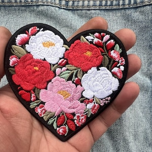 Może przedstawiać: Haftowana naszywka w kształcie serca z kwiatowym wzorem. Naszywka ma czarną obwódkę i zawiera czerwone, białe i różowe kwiaty z zielonymi liśćmi. Naszywka jest trzymana w dłoni osoby na tle dżinsu.