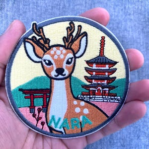 Könnte beinhalten: Runder, gestickter Aufnäher mit einem Hirsch mit Geweih, einer japanischen Pagode und einem roten Torii-Tor. Der Aufnäher hat einen gelben Hintergrund mit dem Wort "NARA" in Grün. Der Hirsch ist braun und weiß mit schwarzen Augen.