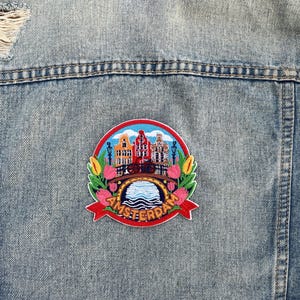 Puede incluir: Un parche bordado redondo que representa el paisaje urbano de &Aacute;msterdam, con edificios, un puente y agua. El parche est&aacute; rodeado de tulipanes y la palabra "Amsterdam" en rojo. Est&aacute; adherido a una chaqueta vaquera azul claro.