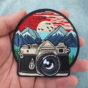 May include: Embroidered patch with a vintage camera design set against a scenic backdrop. The patch shows a sunset with mountains, a lake, and a full moon. The camera is black and beige.