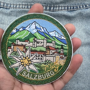 May include: Embroidered patch featuring a scenic view of Salzburg, Austria, with mountains, buildings, and the word "SALZBURG". The patch includes a white flower and musical notes, set against a blue and green background.