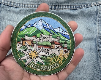 May include: Embroidered patch featuring a scenic view of Salzburg, Austria, with mountains, buildings, and the word "SALZBURG". The patch includes a white flower and musical notes, set against a blue and green background.