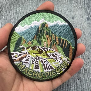 Pode incluir: Remendo bordado circular com uma representação detalhada de Machu Picchu, com montanhas verdes, picos brancos e o texto "MACHU PICCHU". O remendo tem uma borda preta. O remendo é segurado contra um fundo de ganga.