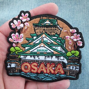 Op de afbeelding: Geborduurde patch met het Osaka-kasteel in Japan, met groene, witte en bruine architectonische details. De patch bevat het woord "OSAKA" in oranje letters en roze kersenbloesems. De patch is zwart langs de randen.