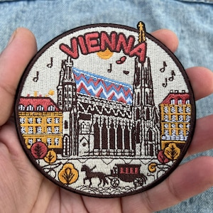 Op de afbeelding: Geborduurde ronde patch met een gedetailleerde illustratie van de Stephansdom in Wenen, met de stadsnaam in het rood. Het ontwerp omvat gebouwen, bomen, muzieknoten en een paardenkoets, alles tegen een beige achtergrond.