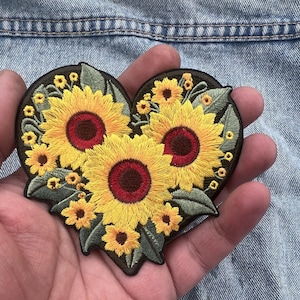 Peut inclure: Écusson brodé en forme de cœur avec des tournesols. Le patch a une bordure marron foncé, des tournesols jaunes avec des centres rouges et des feuilles vertes. Le patch est tenu dans une main.