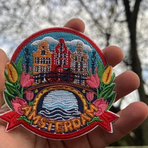 Puede incluir: Parche bordado con un colorido paisaje urbano de &Aacute;msterdam con edificios, un puente y agua. El parche tiene una cinta roja con la palabra "AMSTERDAM" y est&aacute; rodeado de flores y hojas. Un art&iacute;culo de recuerdo o con tem&aacute;tica de viajes.