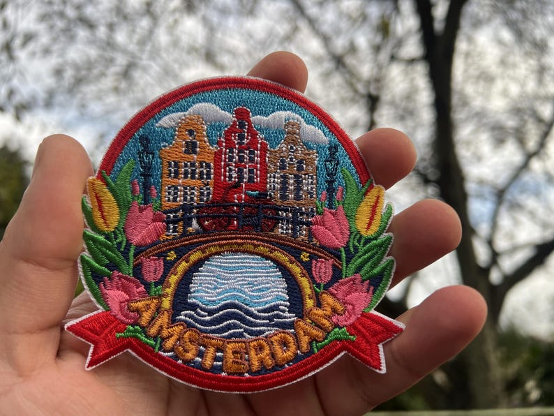 Puede incluir: Parche bordado con un colorido paisaje urbano de &Aacute;msterdam con edificios, un puente y agua. El parche tiene una cinta roja con la palabra "AMSTERDAM" y est&aacute; rodeado de flores y hojas. Un art&iacute;culo de recuerdo o con tem&aacute;tica de viajes.