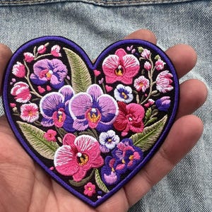 Op de afbeelding: Geborduurde hartvormige patch met een bloemenontwerp. De patch heeft een zwarte achtergrond en een paarse rand. De bloemen zijn roze, paars en wit, met groene bladeren. Voor kleding of accessoires.