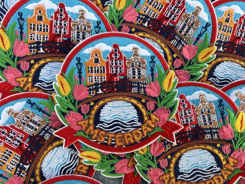 Puede incluir: Parche bordado con un paisaje urbano con tres edificios coloridos, un puente y tulipanes. La palabra "AMSTERDAM" se muestra en una pancarta roja. El parche tiene un fondo de cielo azul y est&aacute; rodeado de hojas verdes.