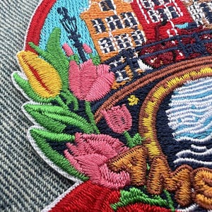 Puede incluir: Parche bordado con un dise&ntilde;o colorido de tulipanes, edificios y un canal. El parche tiene un borde de arco&iacute;ris y la palabra "Amsterdam" en hilo naranja. El parche est&aacute; adherido a una tela vaquera.