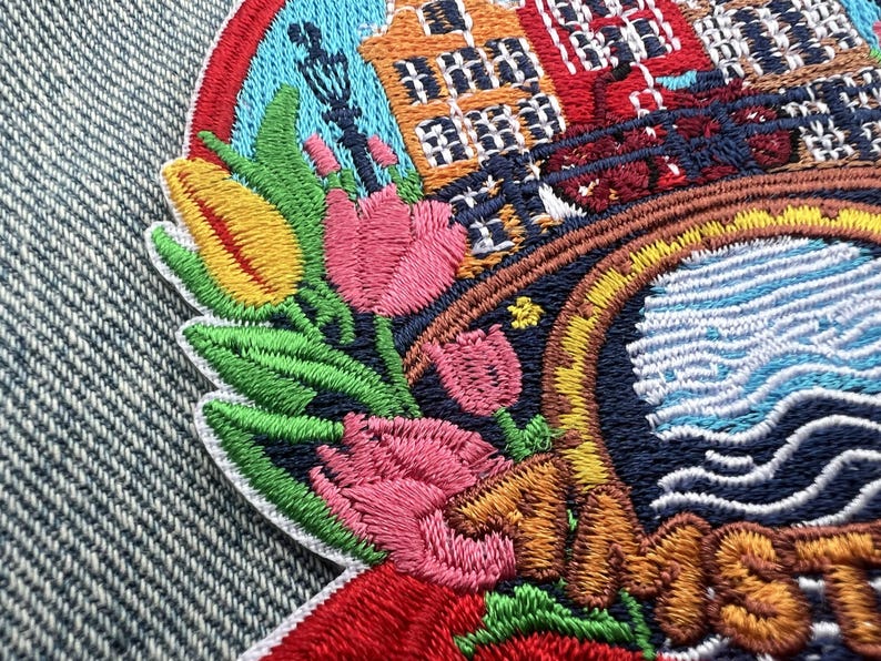 Puede incluir: Parche bordado con un dise&ntilde;o colorido de tulipanes, edificios y un canal. El parche tiene un borde de arco&iacute;ris y la palabra "Amsterdam" en hilo naranja. El parche est&aacute; adherido a una tela vaquera.