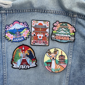 Peut inclure: Cinq écussons brodés représentant des monuments et des villes japonaises sont affichés sur une veste en jean. Les écussons comprennent des images du Mont Fuji, d'une pagode, d'un cerf et des villes de Kyoto, Osaka et Tokyo.