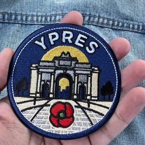 Op de afbeelding: Een ronde geborduurde patch met een donkerblauwe achtergrond en de tekst YPRES in witte letters. Het ontwerp toont een lichtkleurig stenen boogmonument, een gele zon en een rode klaproos centraal op de voorgrond.