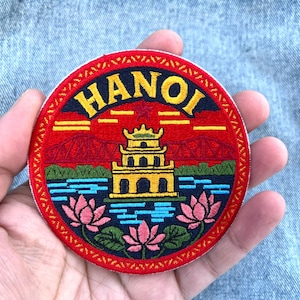 Op de afbeelding: Ronde geborduurde patch met het woord "HANOI" in geel. Het ontwerp toont een pagode, bruggen, water en lotusbloemen op een rode achtergrond. De patch wordt in een hand gehouden.