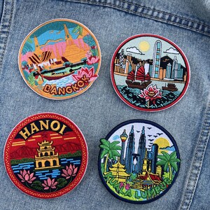 Op de afbeelding: Vier geborduurde patches met reis thema's. De ronde patches tonen scènes uit Bangkok, Hanoi, Kuala Lumpur en een skyline. Elke patch heeft een uniek kleurenschema en ontwerp, met de stadsnaam in tekst.