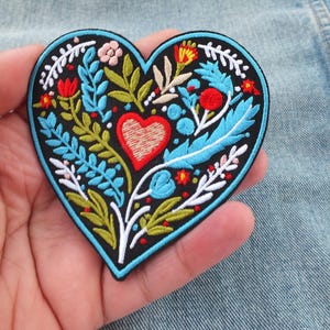 Puede incluir: Parche bordado en forma de corazón con fondo negro y borde azul. El parche presenta un corazón rojo central y está decorado con coloridos diseños florales y de hojas en azul, verde, rojo y blanco. El parche se sostiene en una mano.